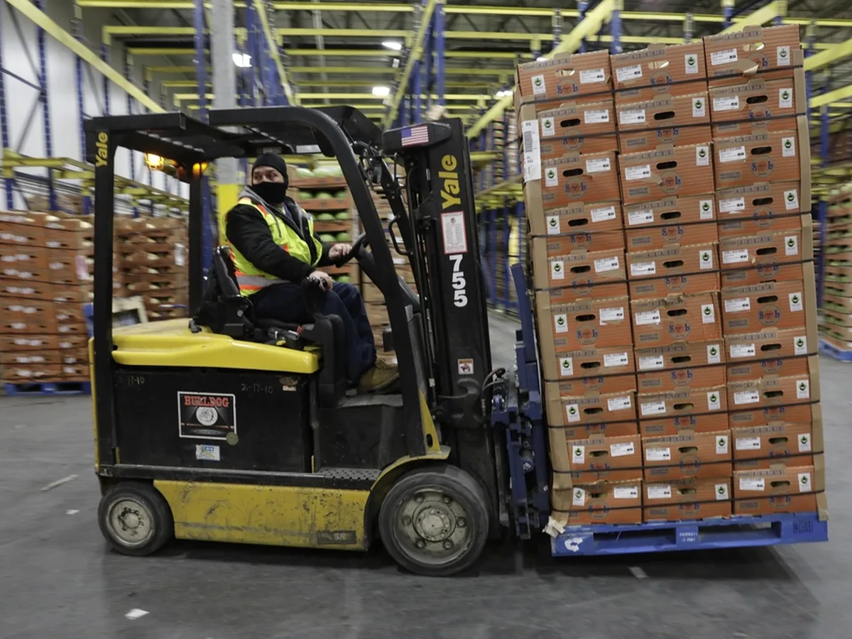 A forklift operator moves a pallet of boxed goods through a distribution warehouse.