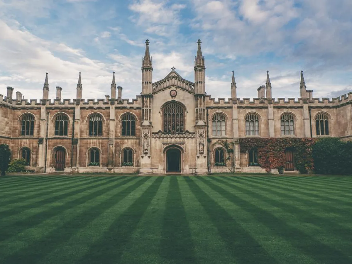 The Gothic front quadrangle of a Cambridge college with a striped lawn under a blue sky.