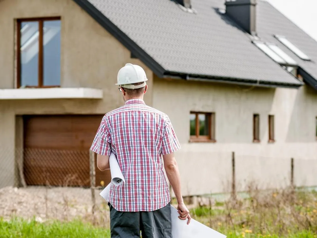 A builder in a hard hat walks towards a newly built house.