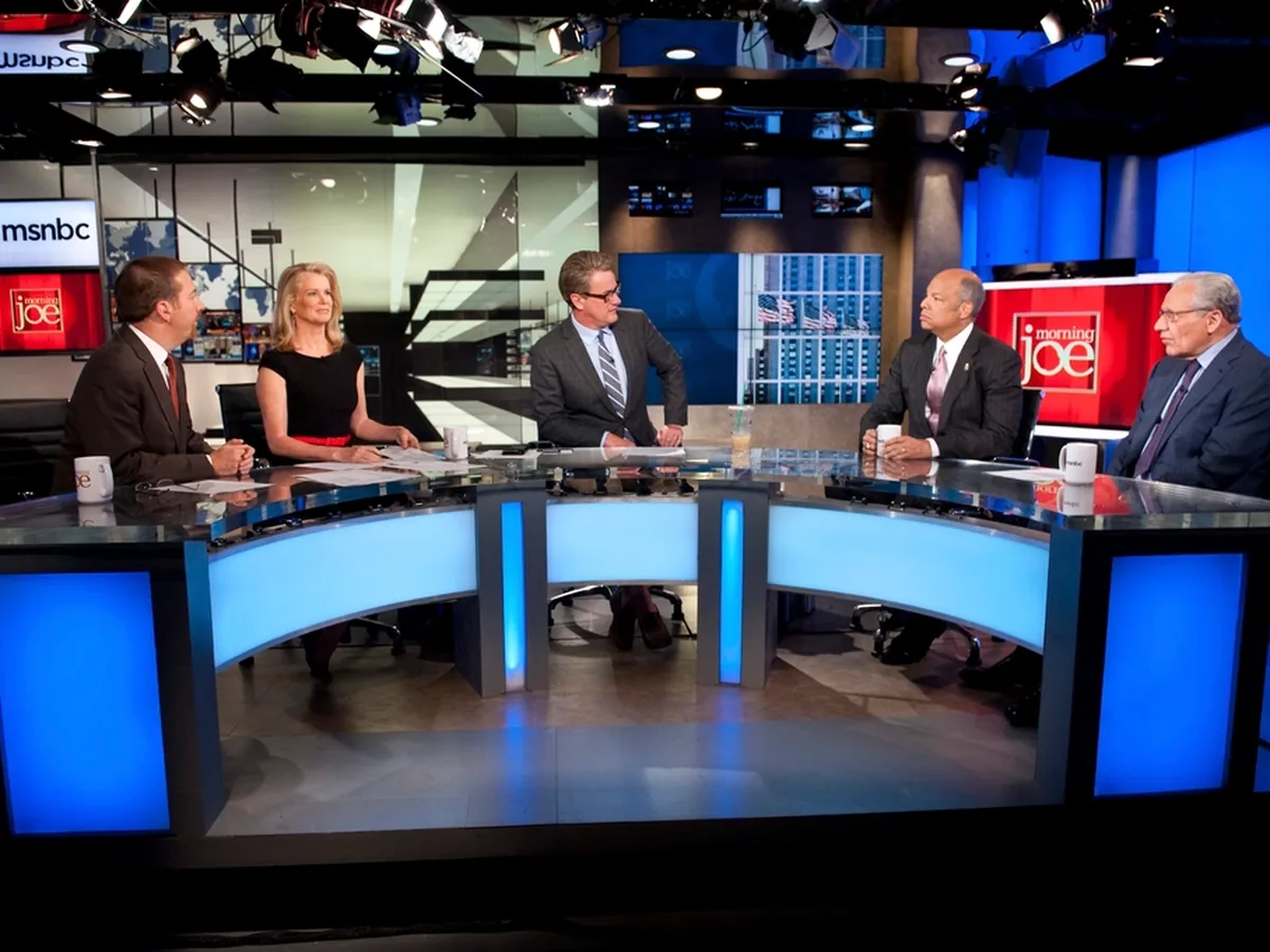 Presenters and guests around the desk of a television news studio.